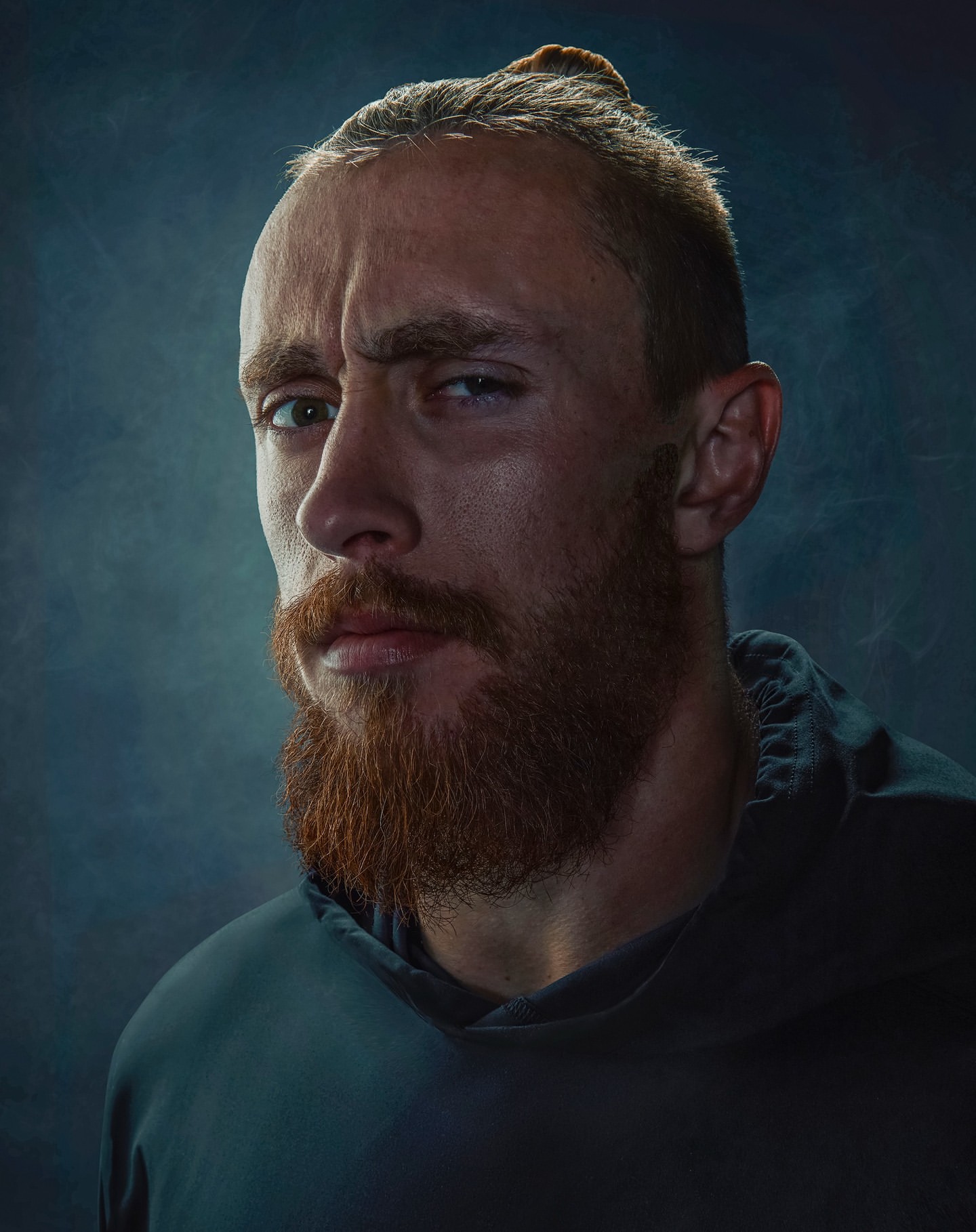 Portrait of a bearded adult male with hair in a bun, looking toward the camera against a dark, slightly textured background with side lighting.
